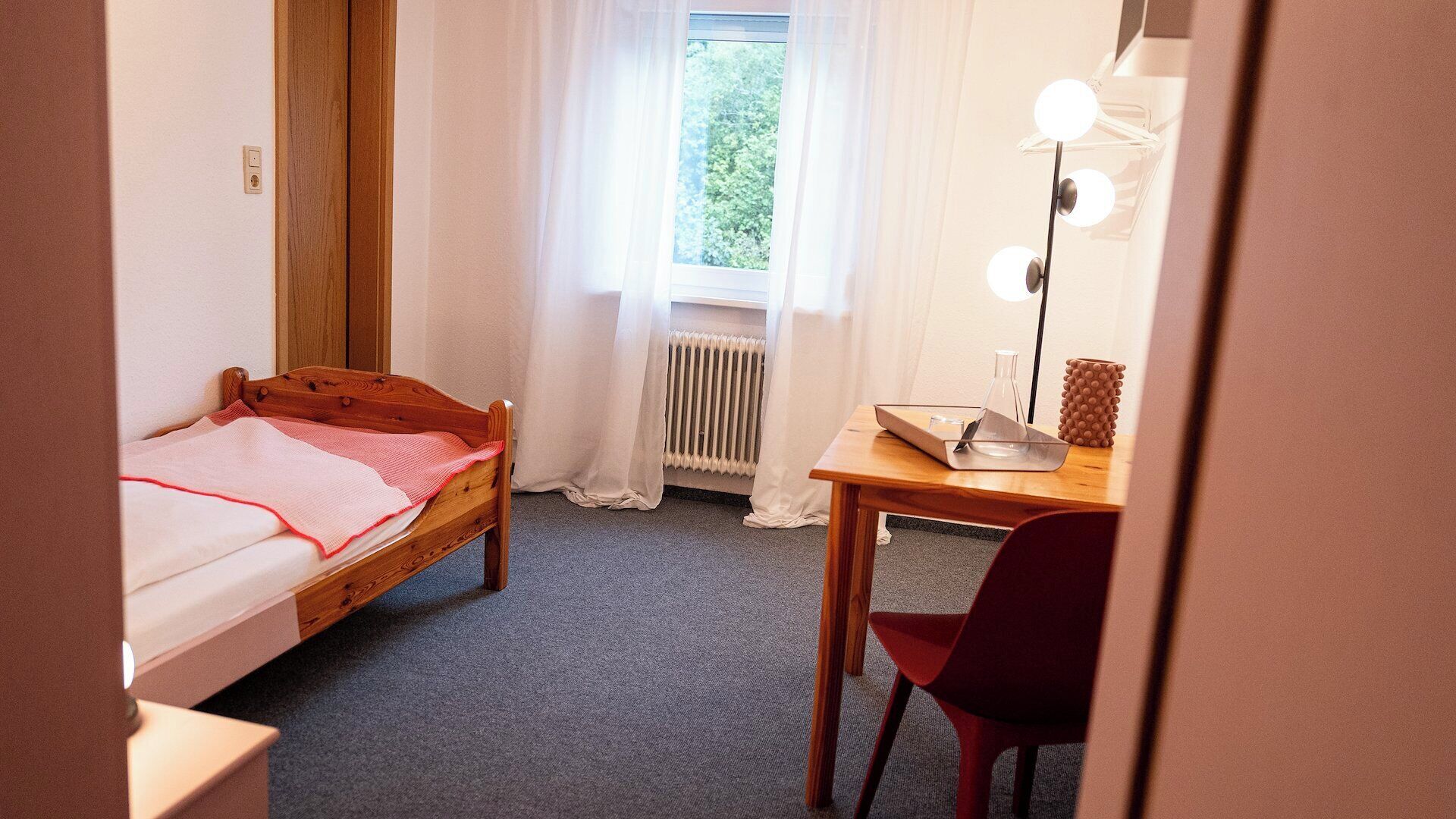 A small, neatly arranged room with a single bed, a desk with a chair, a radiator, and a window with curtains.