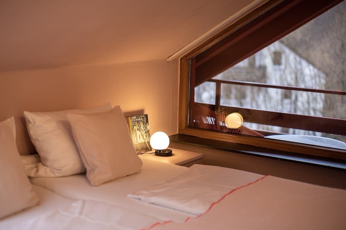 Une chambre à coucher avec un lit, des oreillers, une table de chevet avec une lampe et une fenêtre donnant sur un immeuble.