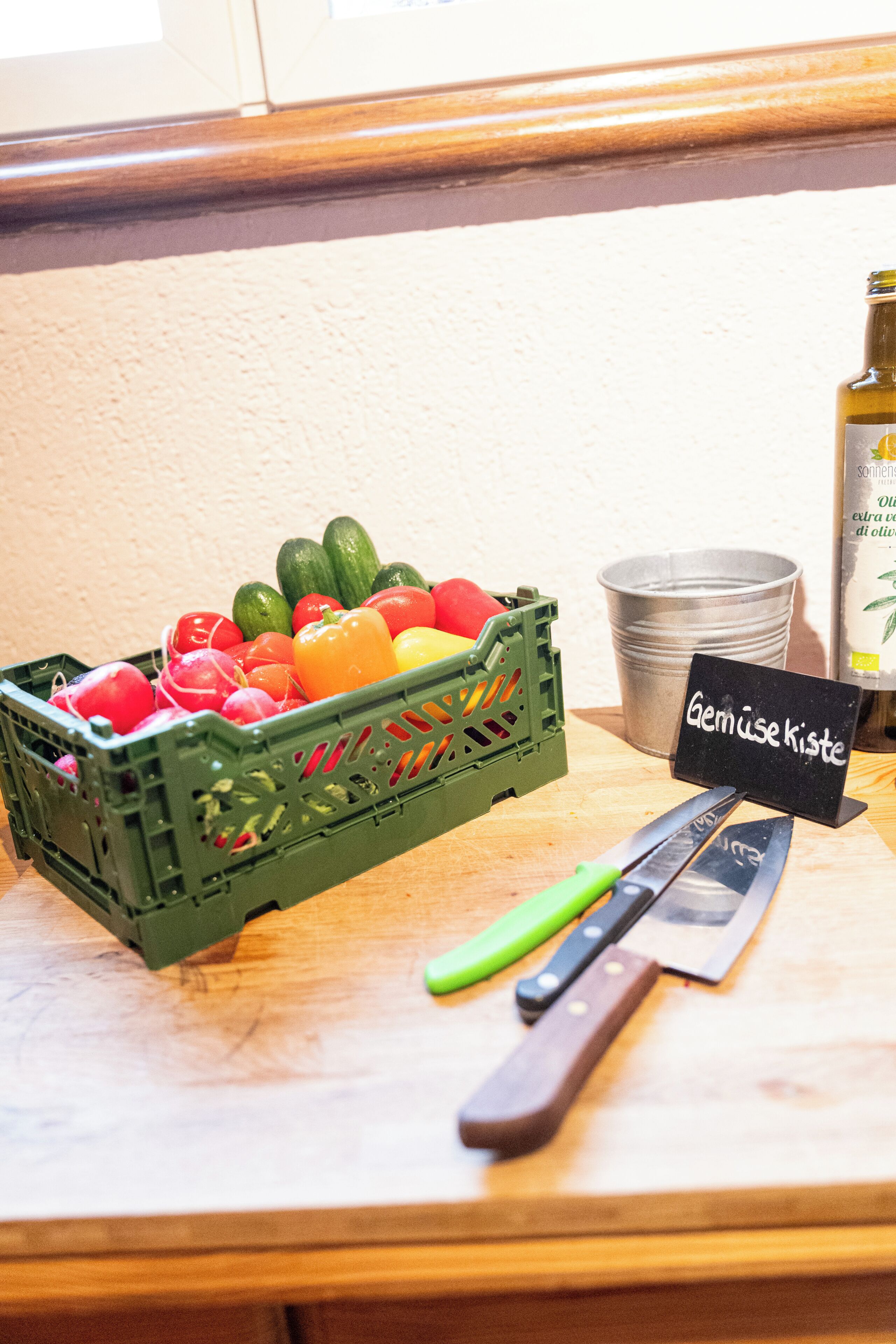A green plastic crate filled with various vegetables, a metal bucket, a bottle of olive oil, and a sign with text.