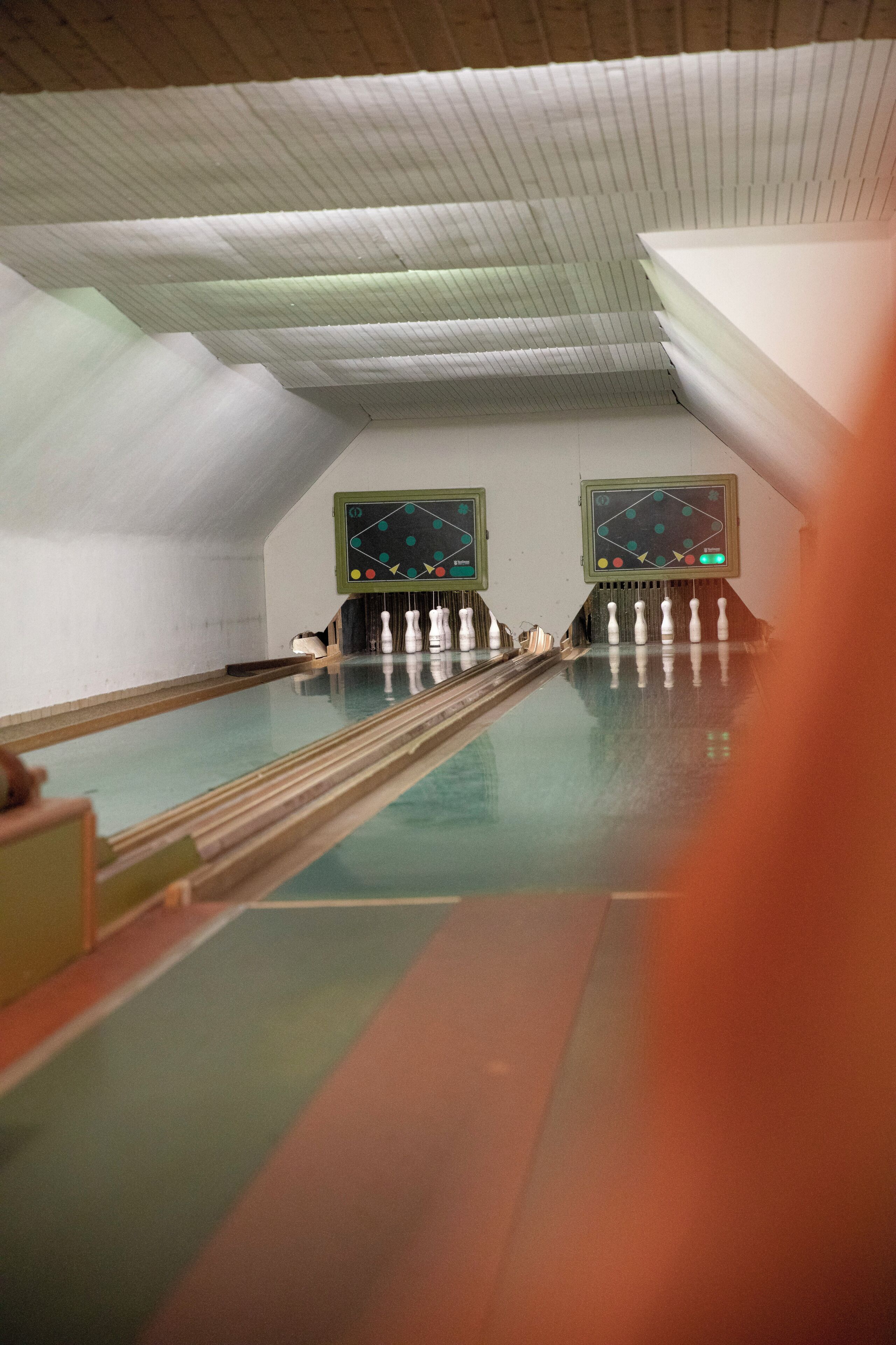 A bowling alley with two bowling lanes, a wooden ceiling, and two scoreboards.