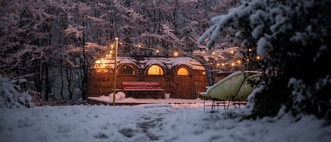 A rustic wooden building with a bench and string lights, surrounded by snow-covered trees.