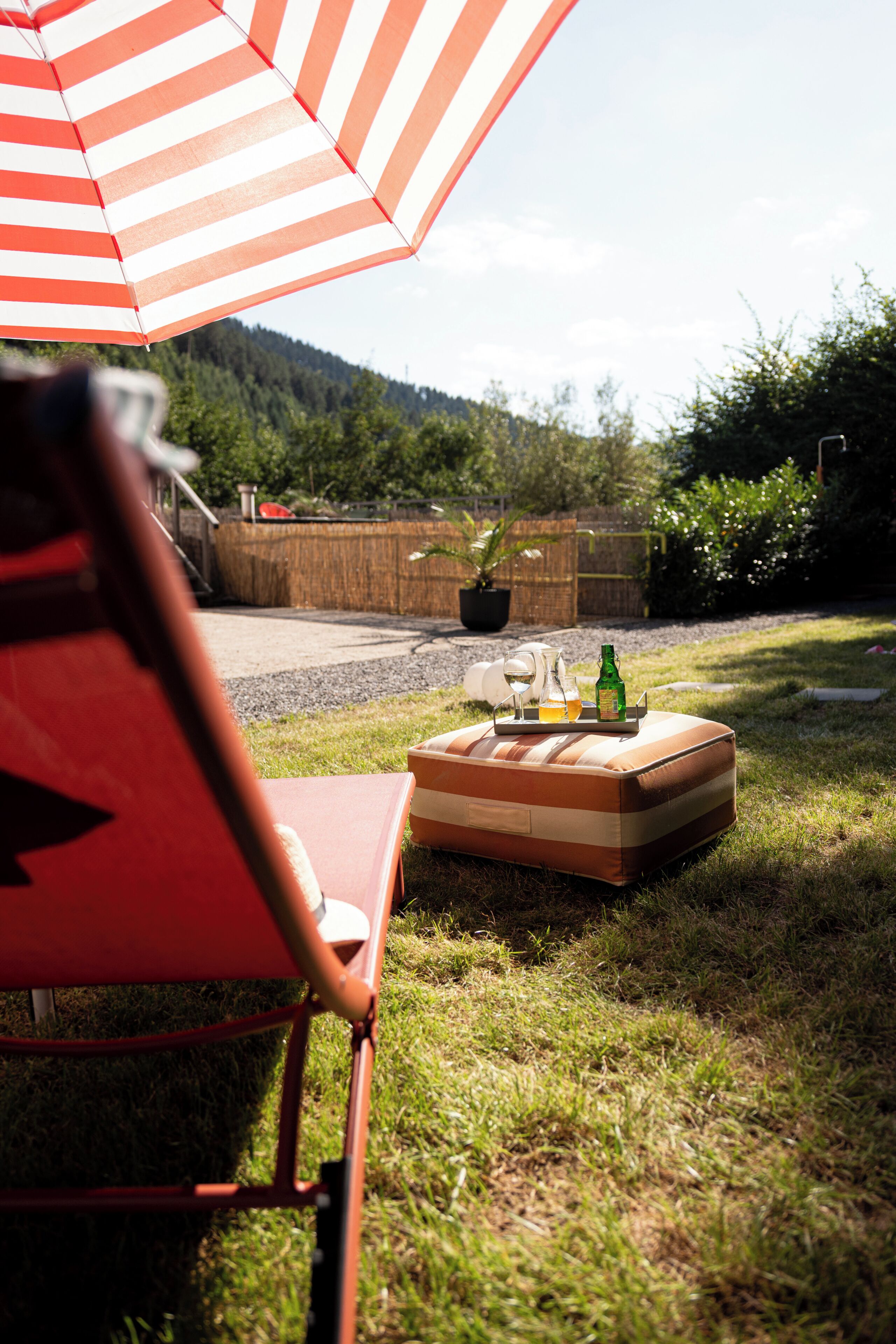 A red chair under a striped umbrella, a wooden bench with bottles and a glass, and a grassy area with a mountain in the background.