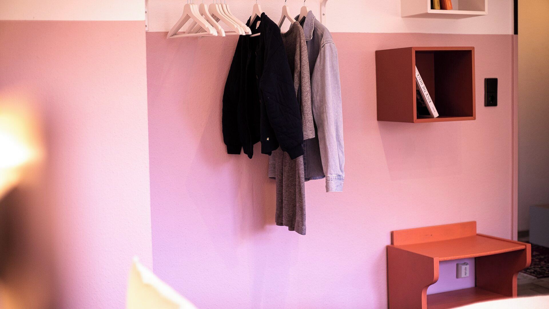 A room with a pink wall, a wooden hanger rack with clothes, a small wooden shelf, and a red bench.