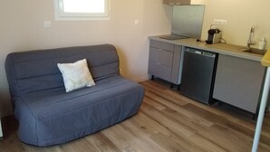 Un espacio habitable funcional con un sofá gris, una pequeña cocina que incluye lavavajillas, y un ventanal que proporciona luz natural.