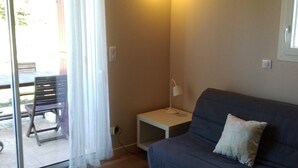 Una habitación con un sofá azul oscuro, una mesita de noche blanca pequeña con una lámpara, y un ventanal corredero que da a un espacio exterior con una mesa y sillas.