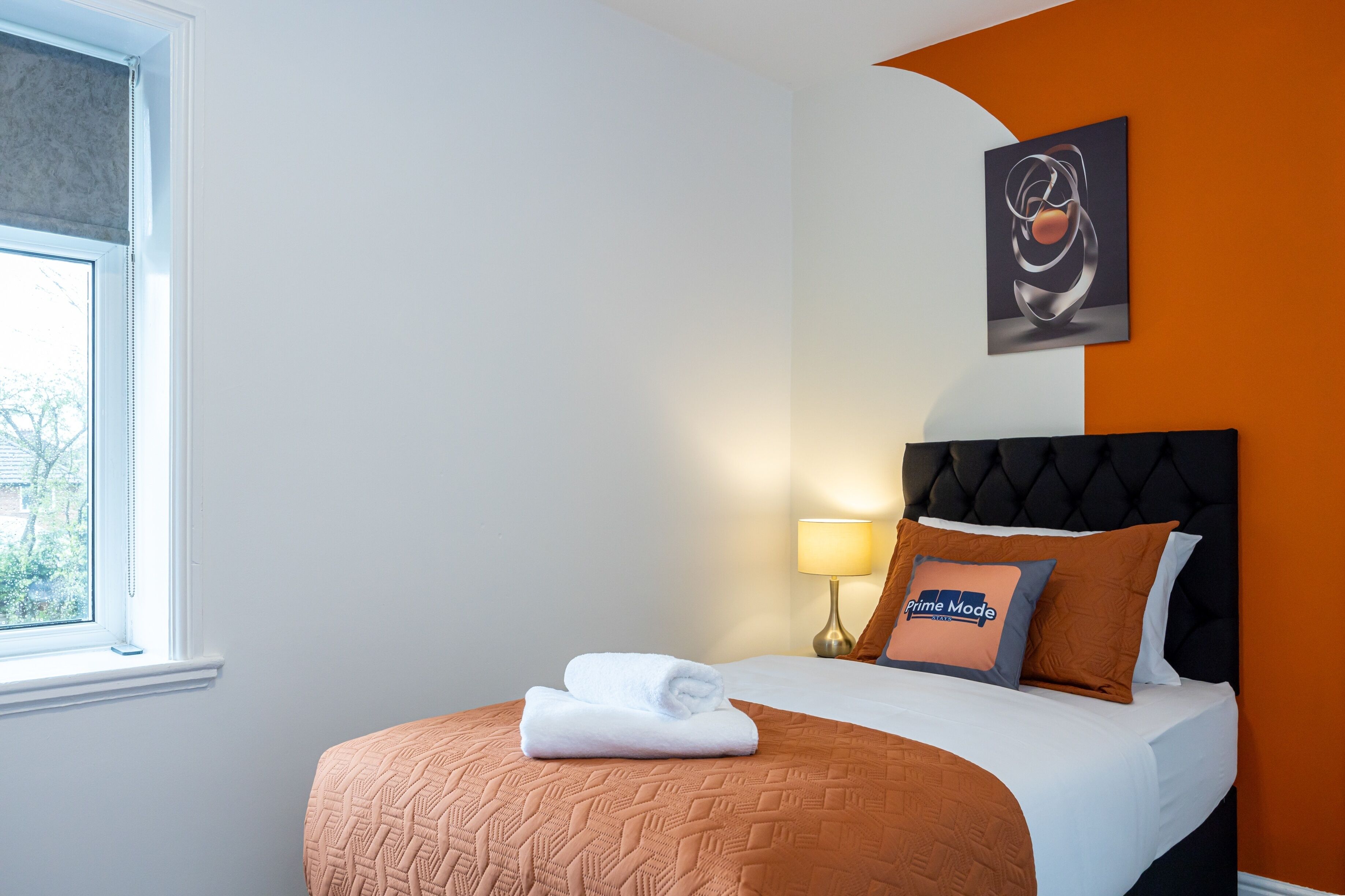 A hotel room with a bed, a window, a wall-mounted lamp, and a painting.