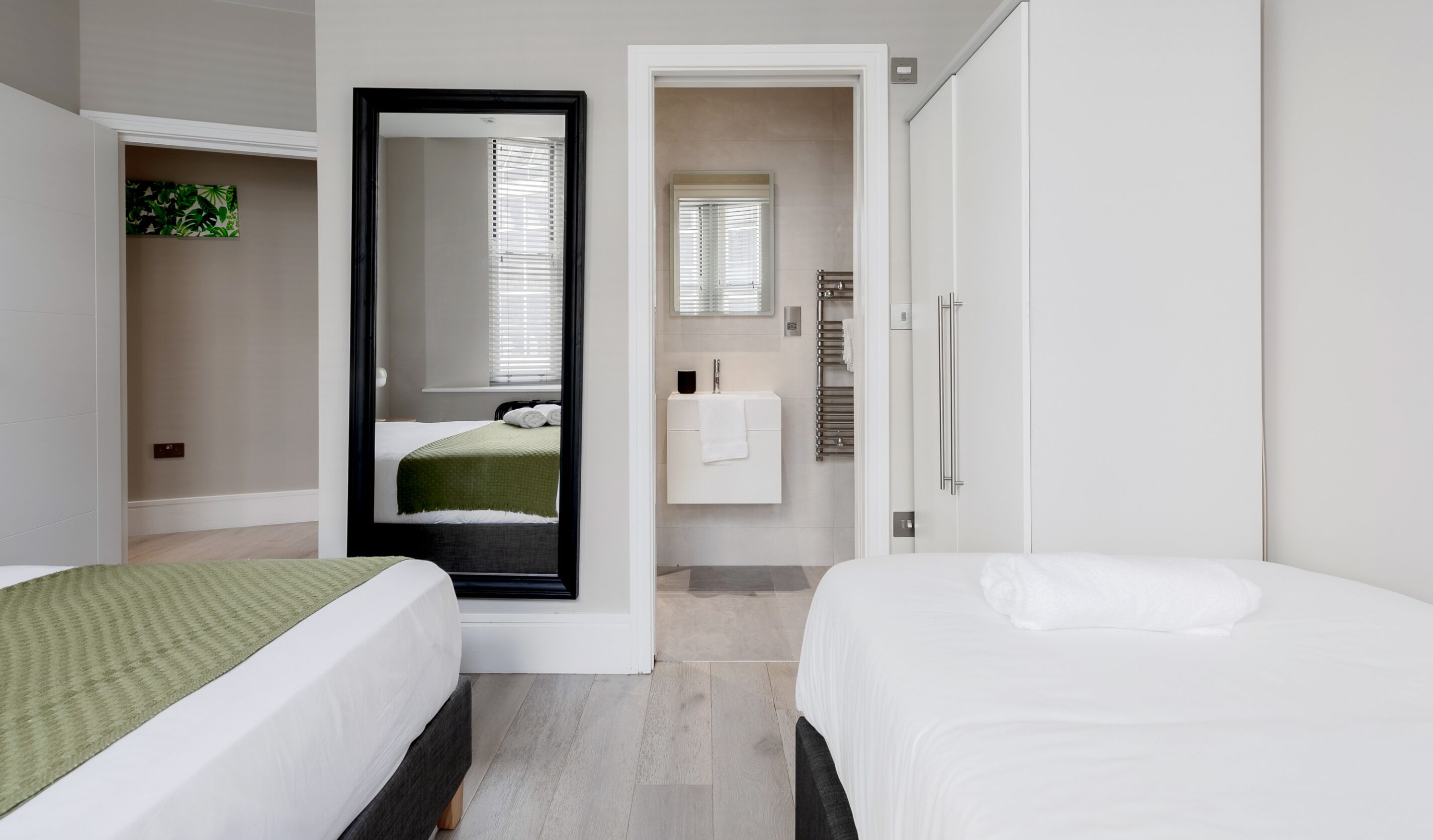Una camera d'albergo con due letti, un bagno con lavandino e porta asciugamani, e uno specchio che riflette la stanza.