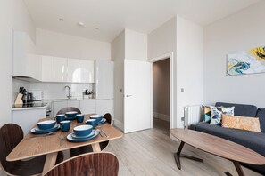 A modern kitchen with white cabinets, a dining table set with blue dishes, and a grey sofa with patterned cushions. - Superb Apartments opp Edgware Rd Station (London)