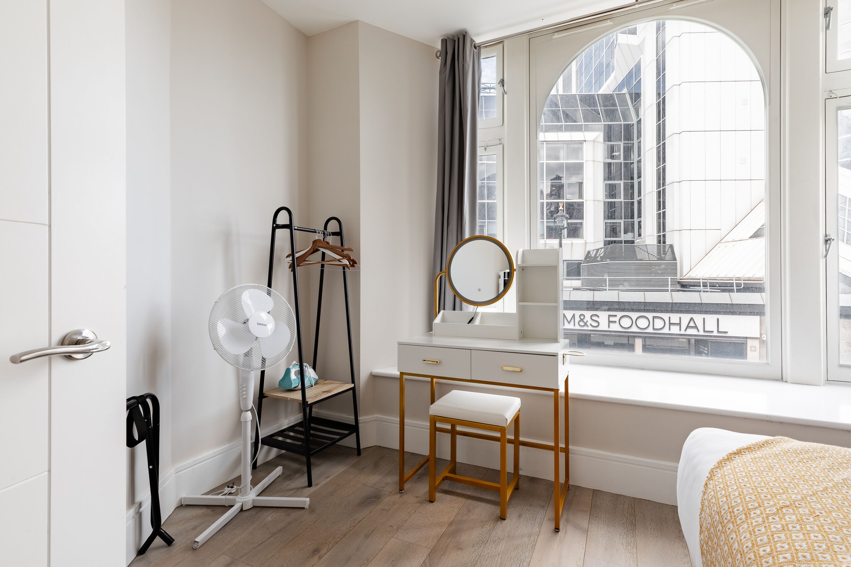 A modern hotel room with a white dresser, a wooden chair, a standing fan, and a window with a view of a building labeled "M&S FOODHALL".