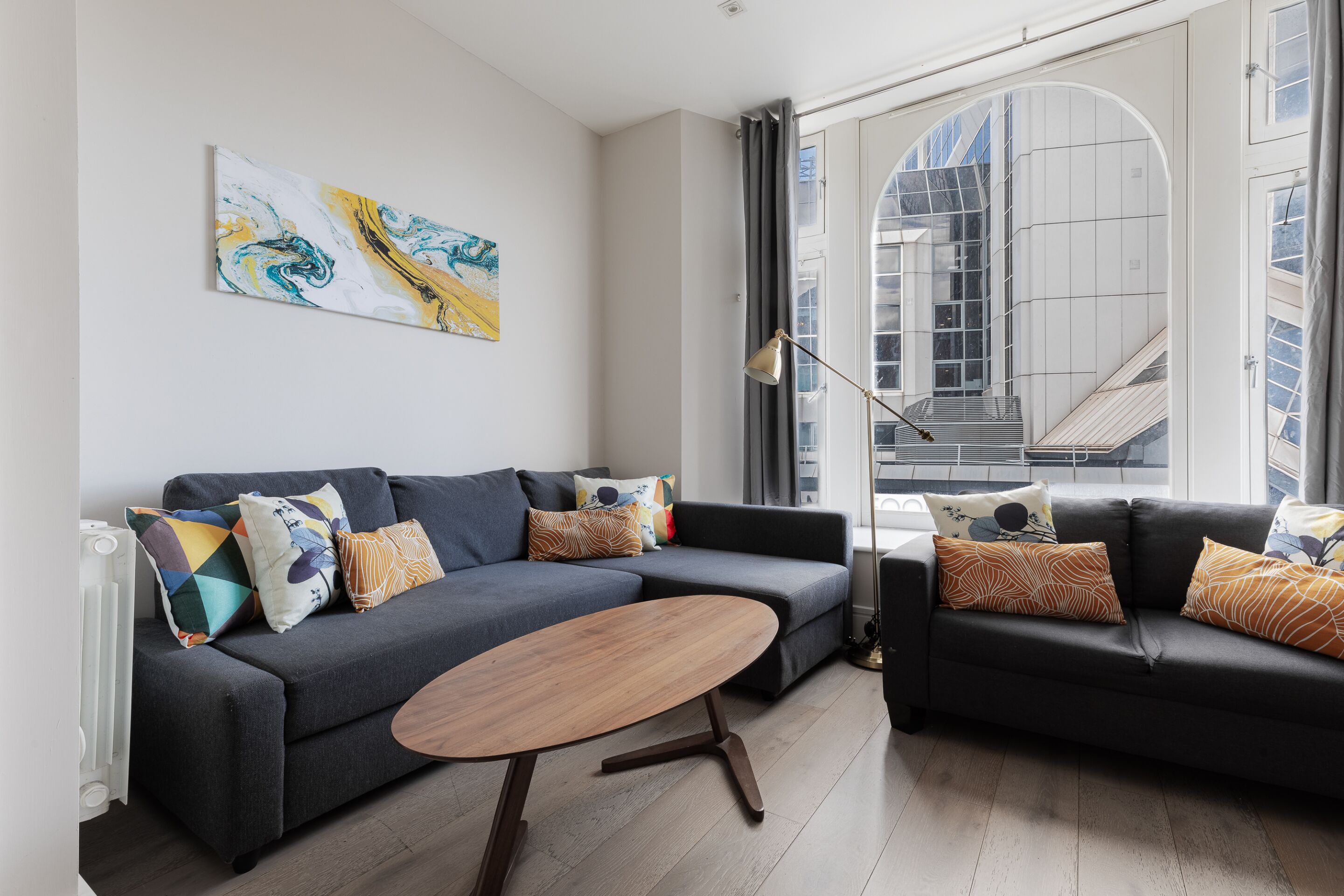 A modern living room with a grey sectional sofa, a wooden coffee table, and a large window with a view of a building.
