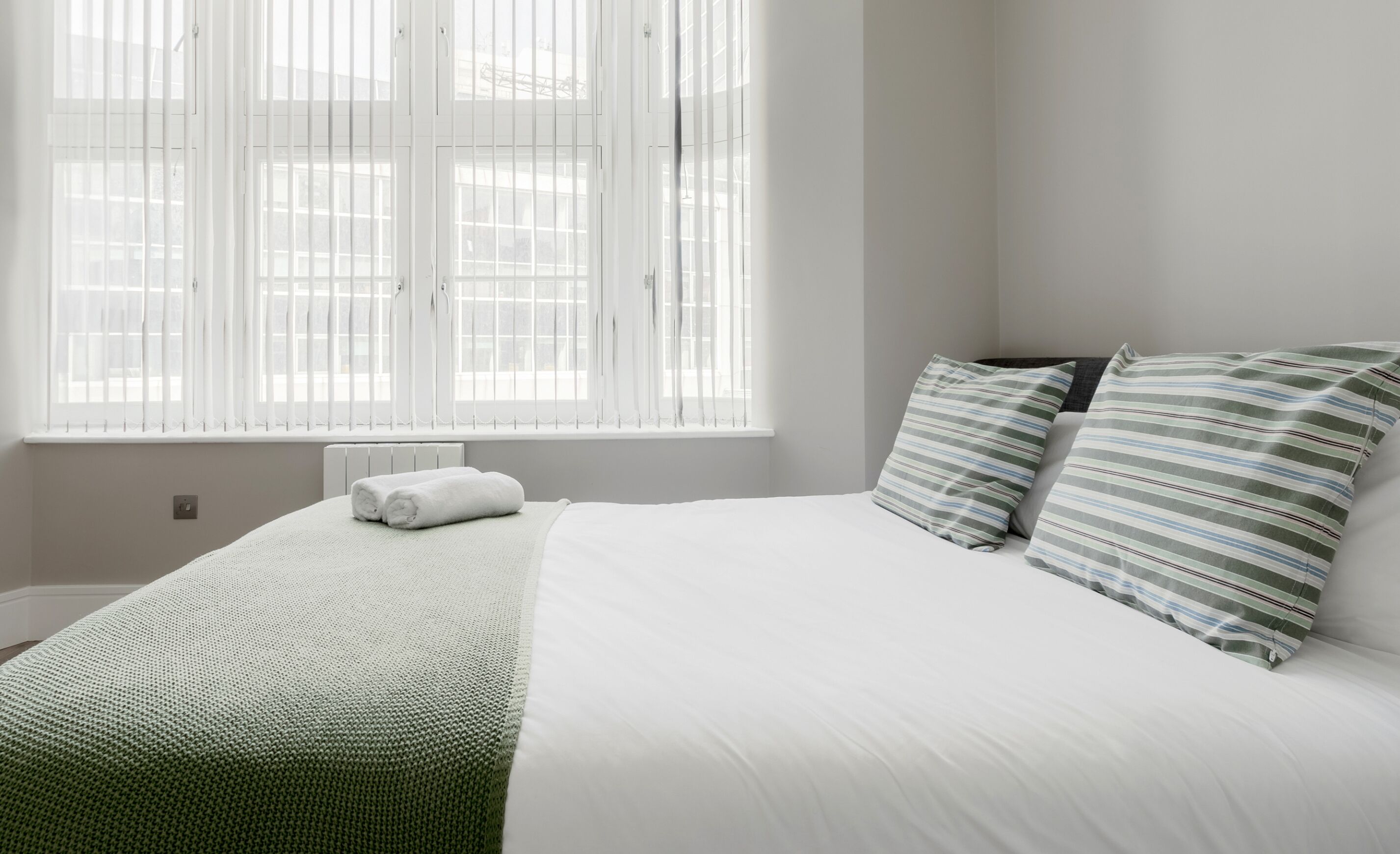 Quarto com cama, travesseiros, janela com persianas e radiador.