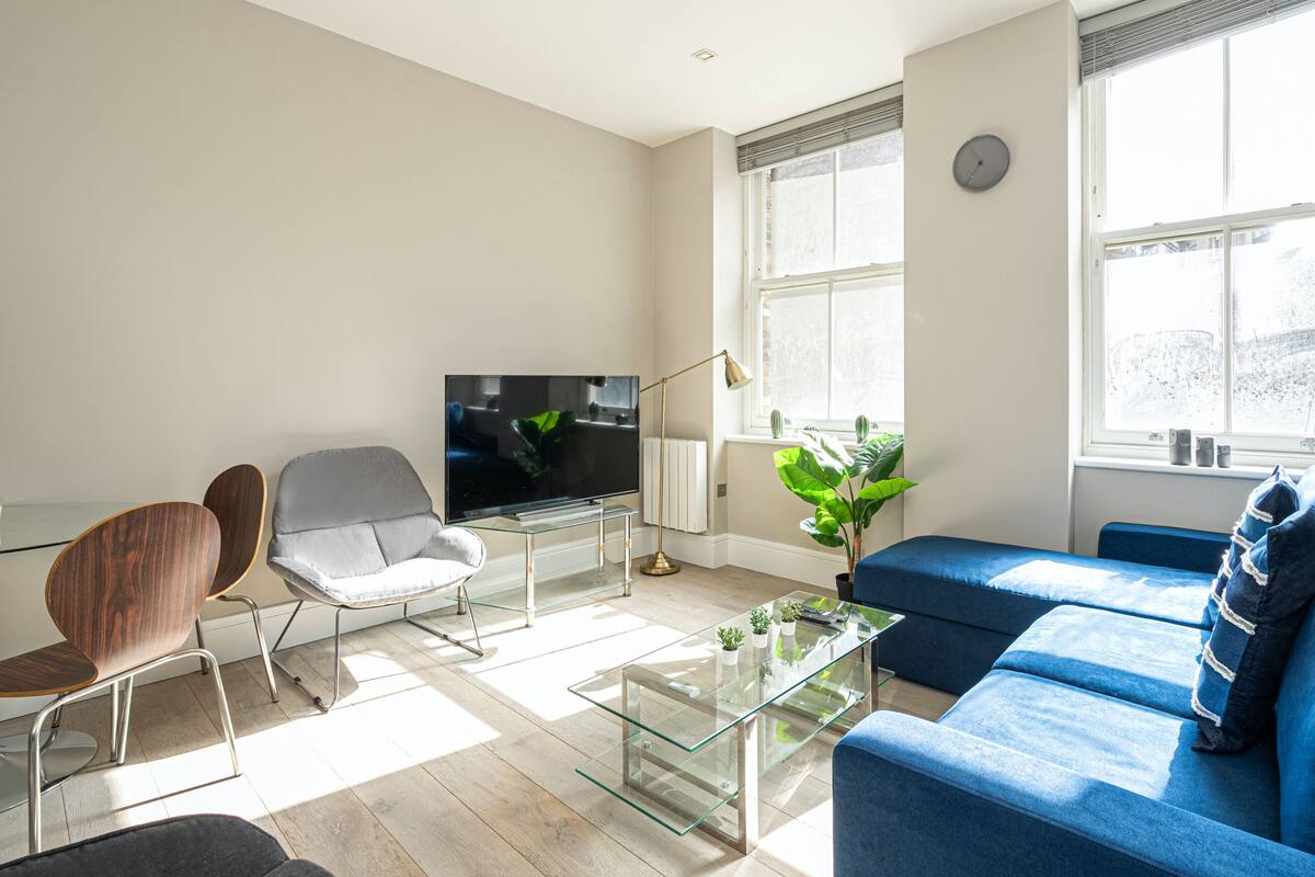 Un salon moderne avec une table basse en verre, un téléviseur à écran plat, un canapé bleu et une grande fenêtre équipée de stores.