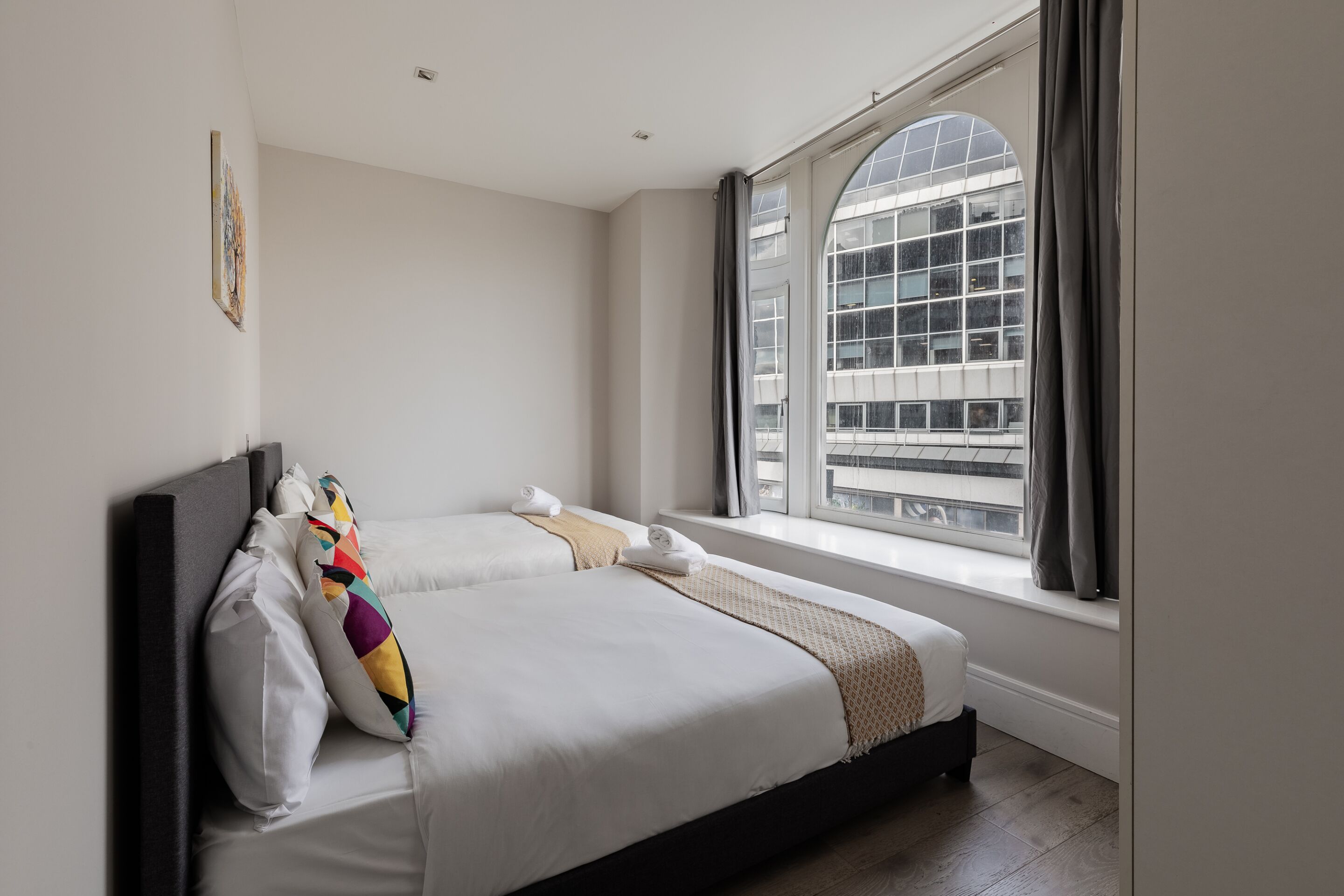 A modern hotel room with a large window, a bed with white linens, a dark headboard, and a colorful pillow.
