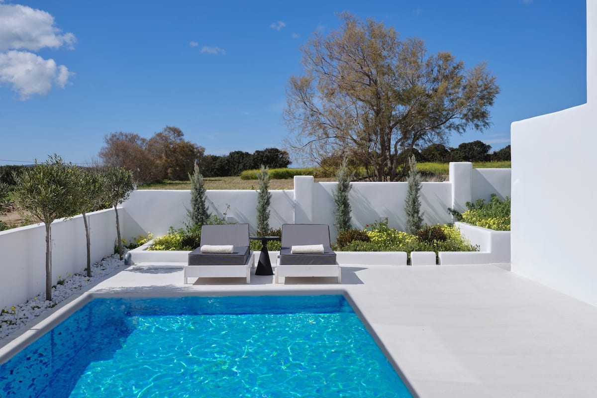 Un área de piscina moderna al aire libre con una piscina azul cristalina, paredes blancas y vistas al campo.