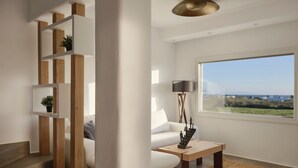 A modern living room with a white sofa, a wooden coffee table, a floor lamp, and a large window offering a view of the outdoors. - Maestro Villas Naxos (Naxos)
