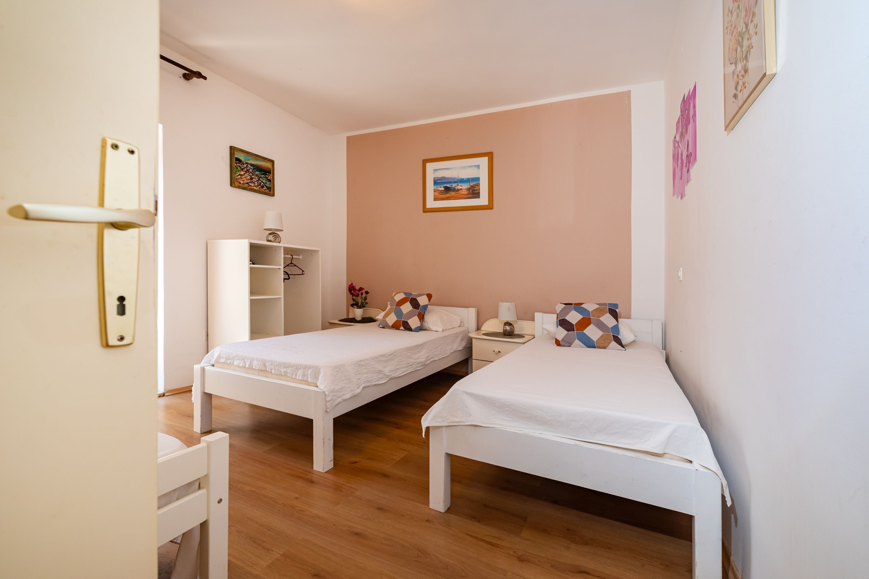 A bedroom with two single beds, a white wardrobe, a small table with a lamp, and a painting on the wall.