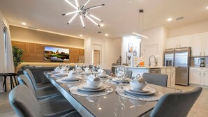 A modern dining area with a long table set for a meal, a kitchen with stainless steel appliances, and a TV mounted on the wall.