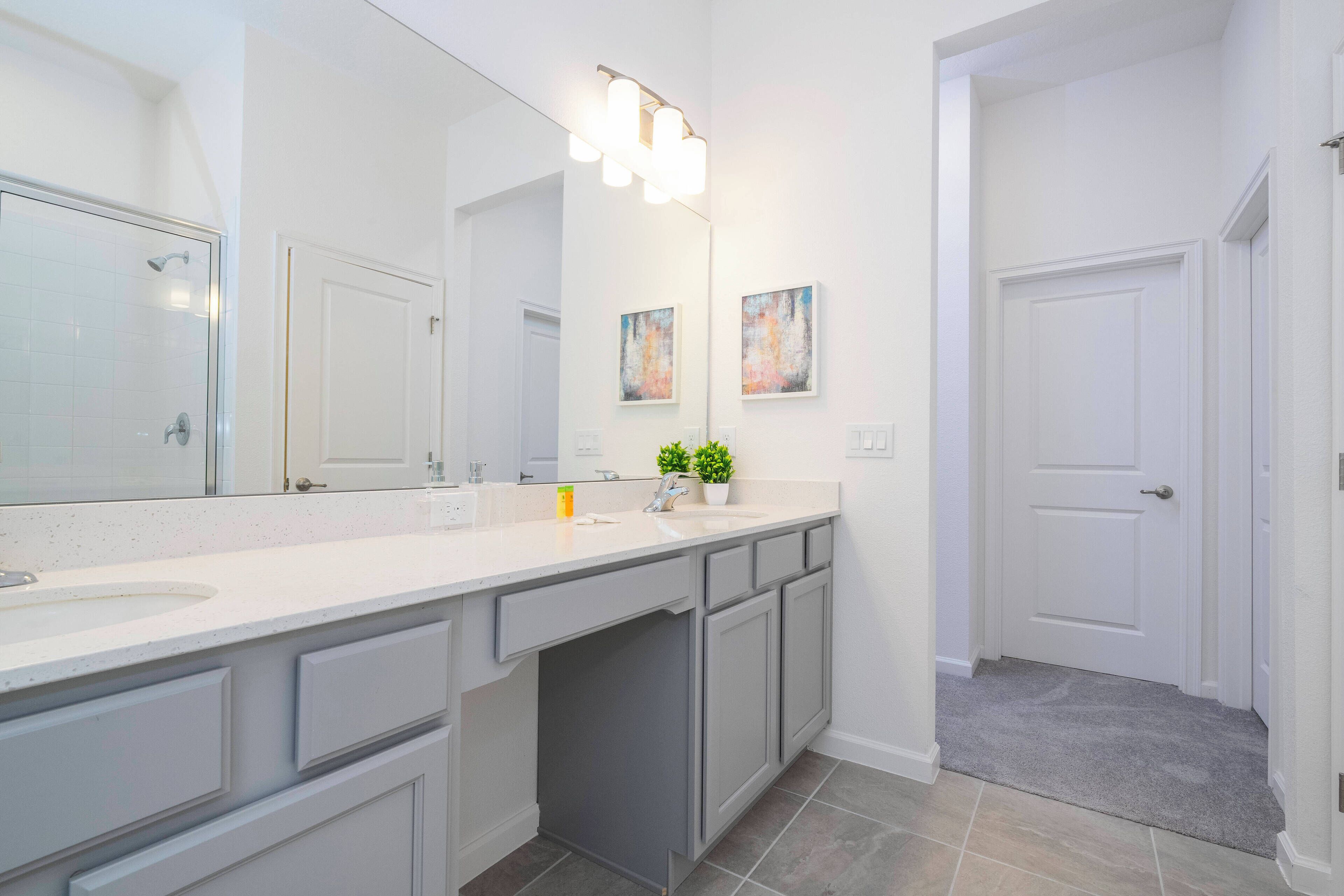 Een moderne badkamer met een grote spiegel, een wastafel en een douche. Twee kunstwerken in lijsten hangen aan de muur en op het aanrecht staat een kleine plant in pot.