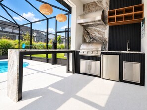 An outdoor kitchen with a built-in grill, a marble backsplash, and a pool in the background.