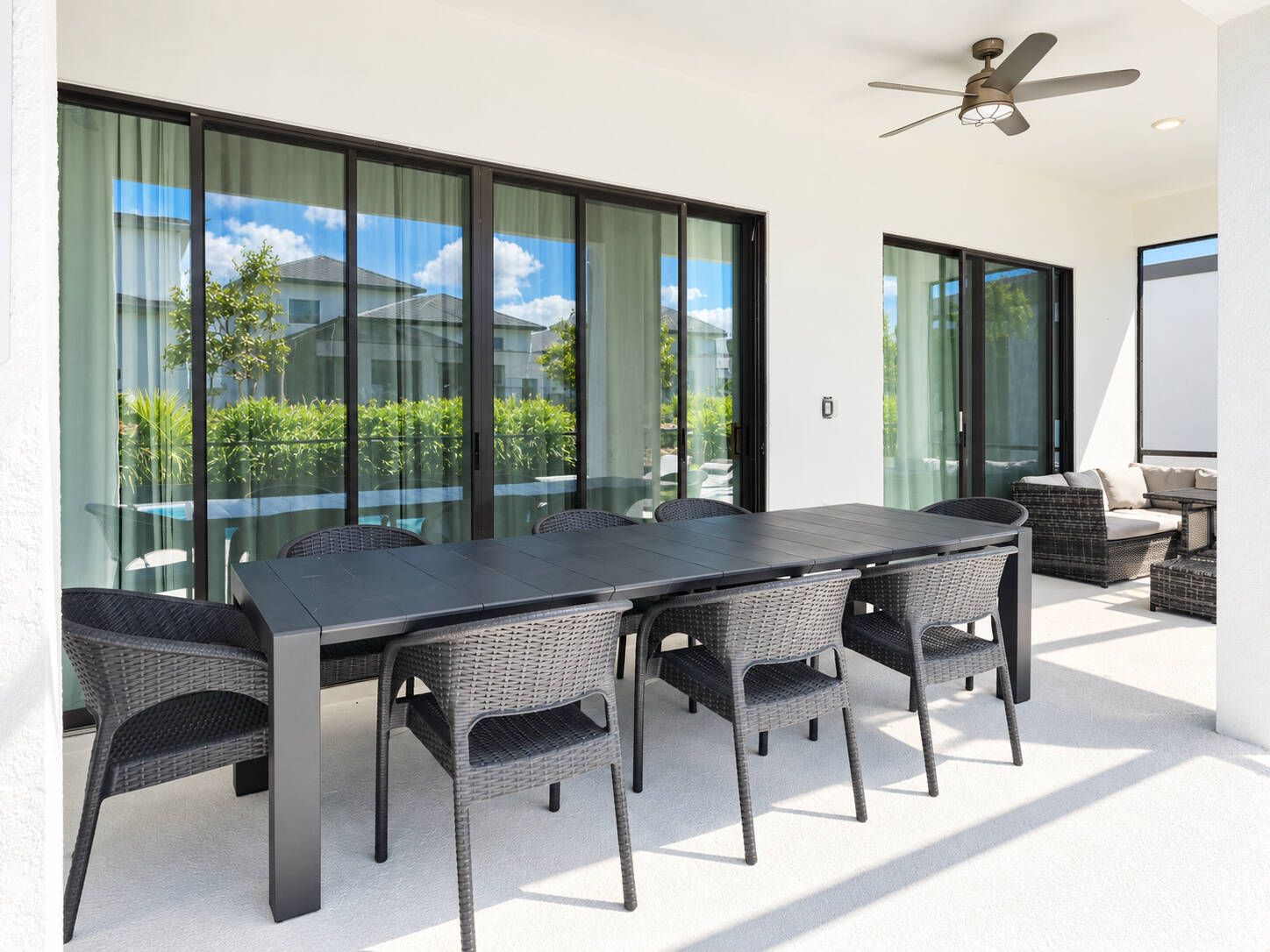 Un espace de restauration extérieur moderne, doté d’une grande table et de chaises, d’un ventilateur de plafond et offrant une vue sur la verdure grâce à de grandes portes vitrées.