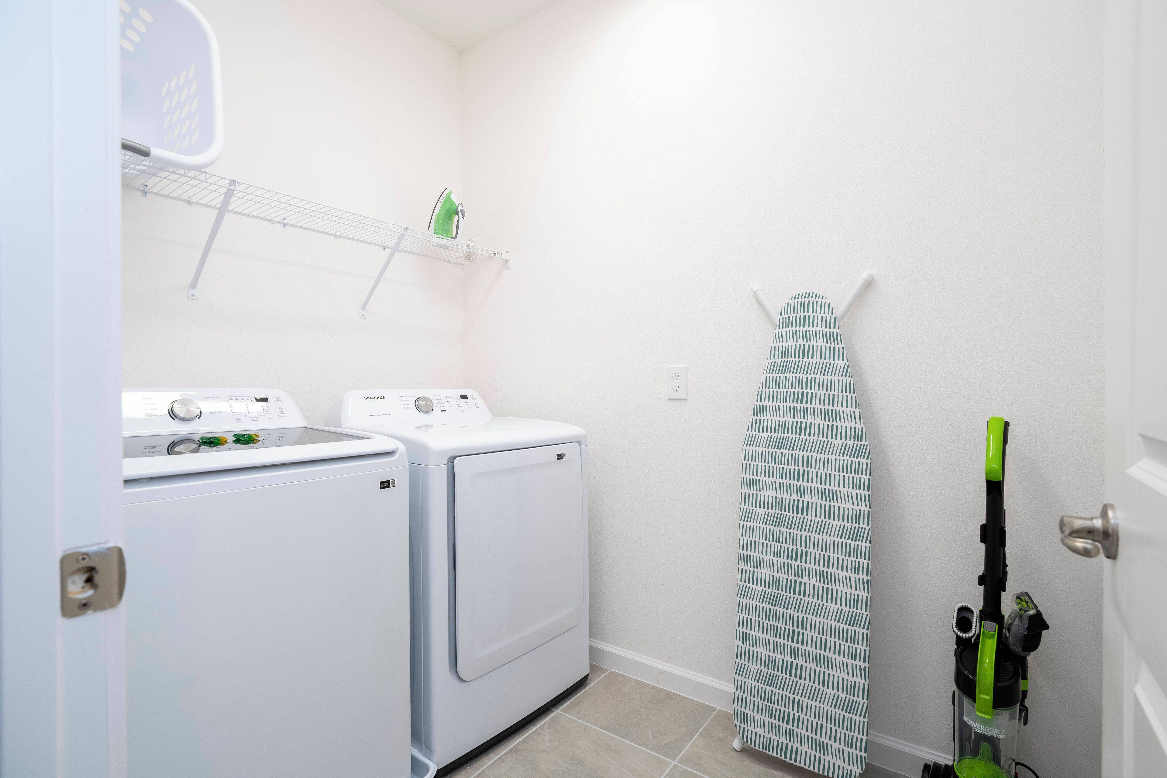 A laundry room with two white washing machines, a drying rack, and a green ironing board.