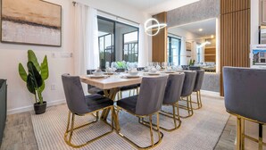 A modern dining area with a large table set for six, surrounded by grey upholstered chairs with gold legs. - The Best Home and Location Near Disney (6139PG) (Orlando)