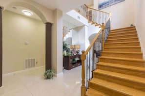 A modern interior with a wooden staircase, a hallway leading to a living area, and a large mirror reflecting the space.