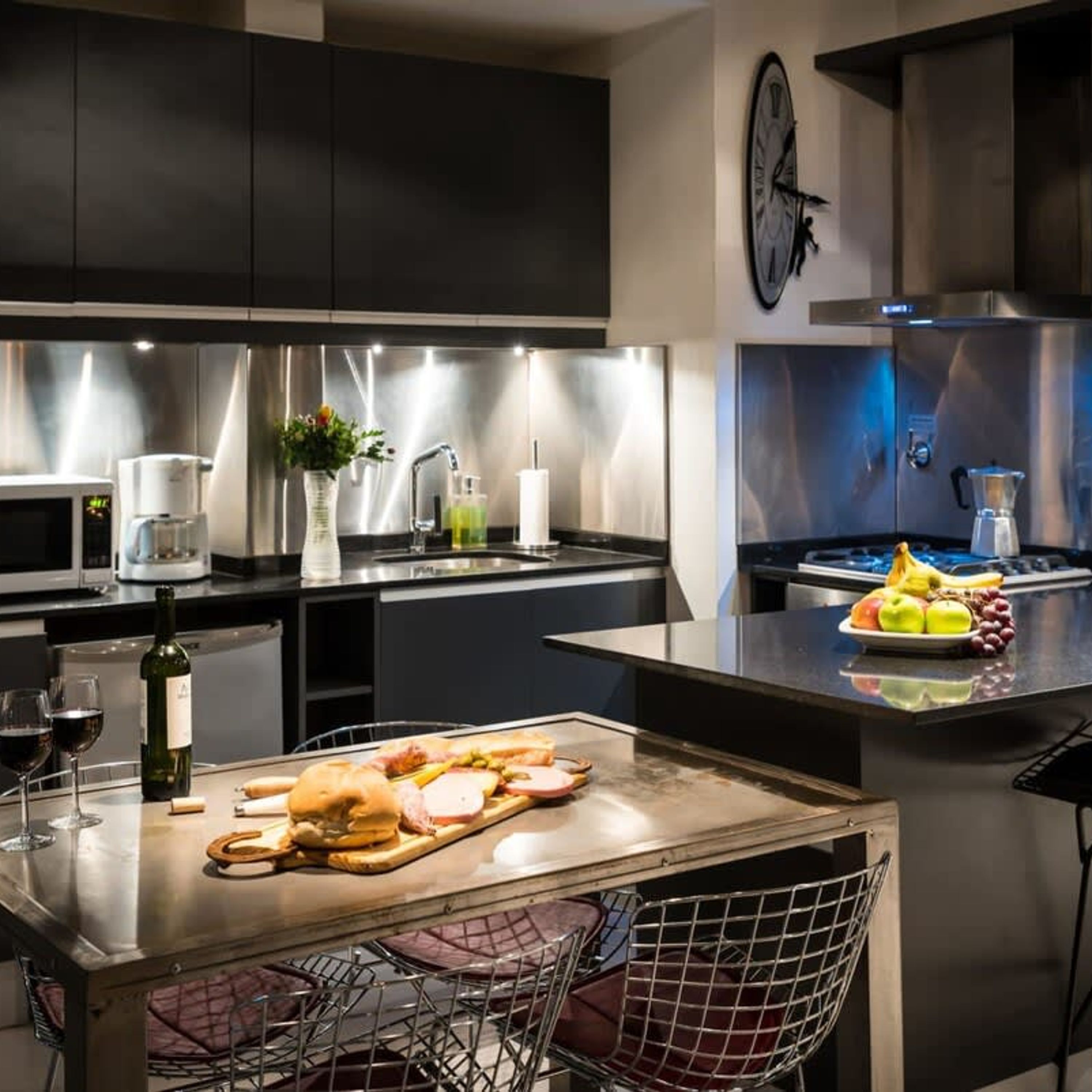 A modern kitchen with dark cabinetry, stainless steel appliances, a clock on the wall, and a fruit bowl on the counter.