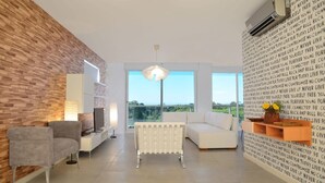 A modern living room with a brick wall, a white radiator, a grey sofa, a white armchair, a TV, and a large window with a view. - Green Bliss Spacious Studio in Nu ez (Buenos Aires)