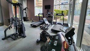 Un gimnasio con máquinas de cardio, equipo de pesas y grandes ventanales con vistas a una zona residencial.
