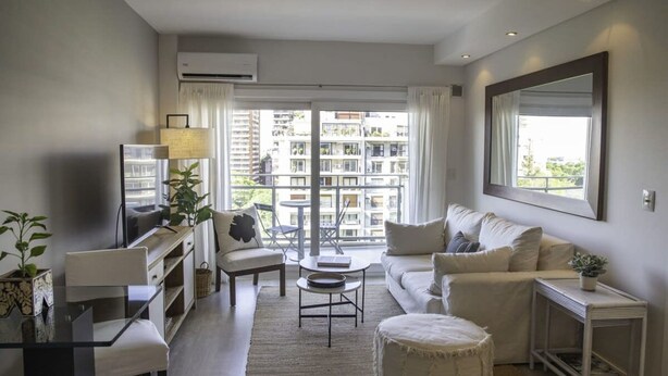Un salon moderne avec un canapé, une table basse et un meuble pour la télévision. Il y a une grande fenêtre avec des rideaux, une plante en pot et un miroir au mur.