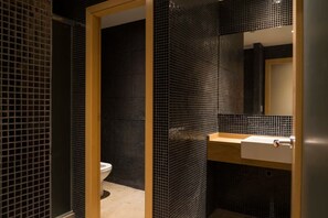 Un baño moderno con pared de azulejos negros, puerta de madera, lavabo blanco y espejo.