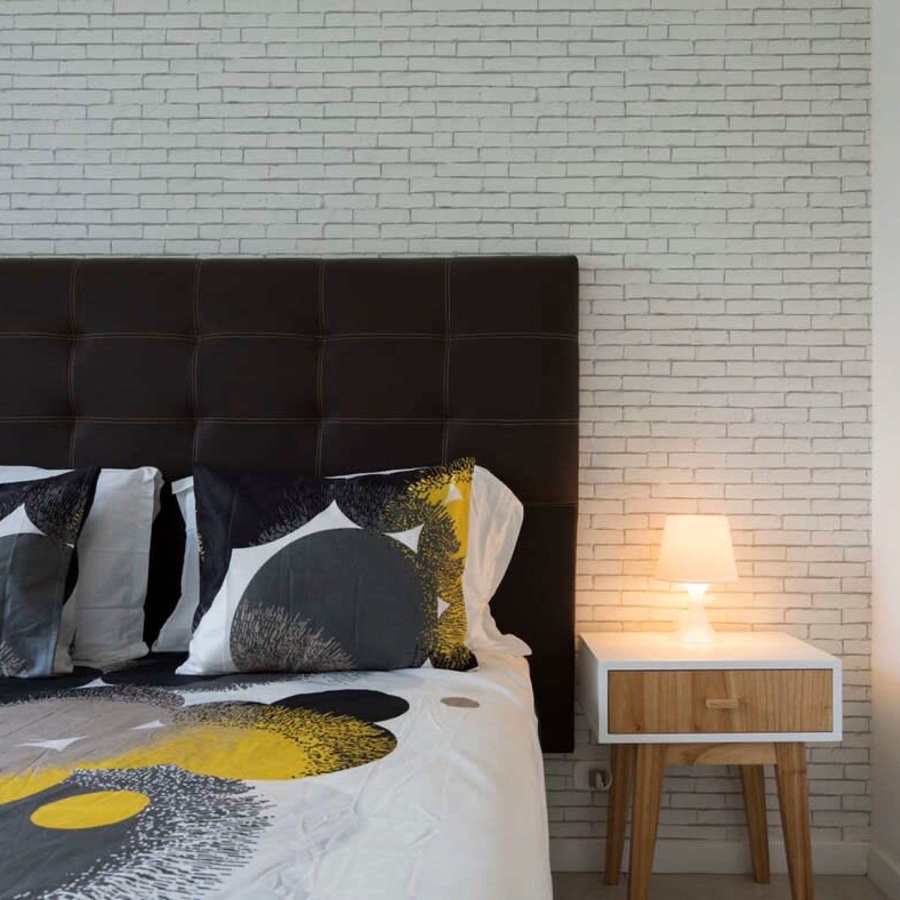 A bed with a dark headboard, a white nightstand with a lamp, and a white brick wall.