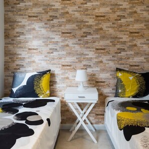 Habitación de hotel con dos camas, cada una con una colcha de estampado blanco y negro, una mesita de noche blanca con una lámpara, y una pared con diseño de ladrillo.