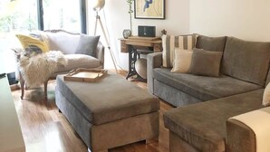 Ein modernes Wohnzimmer mit einem grauen Sofa, einem kleinen Hocker, einem Schreibtisch mit Lampe und einem gerahmten abstrakten Gemälde an der Wand.