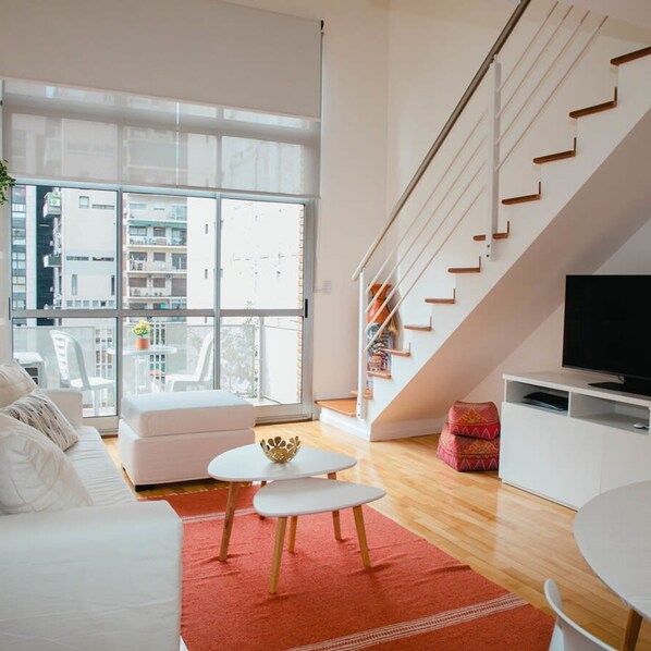 A modern living room with a white sofa, a small white table, a red rug, a staircase with wooden steps, and a TV on a white cabinet.