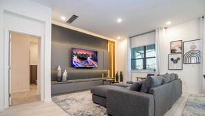 A modern living room with a grey sofa, a flat-screen TV mounted on the wall, and a large window with curtains. - 8BR Made for Families, Built for Fun (5878PG) (Orlando)
