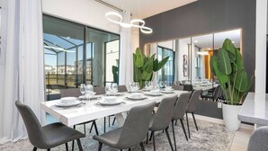 A modern dining area with a long table set for six, surrounded by grey chairs, large green plants, and a white ceiling light fixture. - 8BR Made for Families, Built for Fun (5878PG) (Orlando)