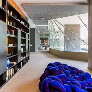 Ein modernes Interieur mit einer großen Bücherwand, einem blauen Sitzsack und Blick auf eine Brücke und ein Gebäude durch große Fenster.