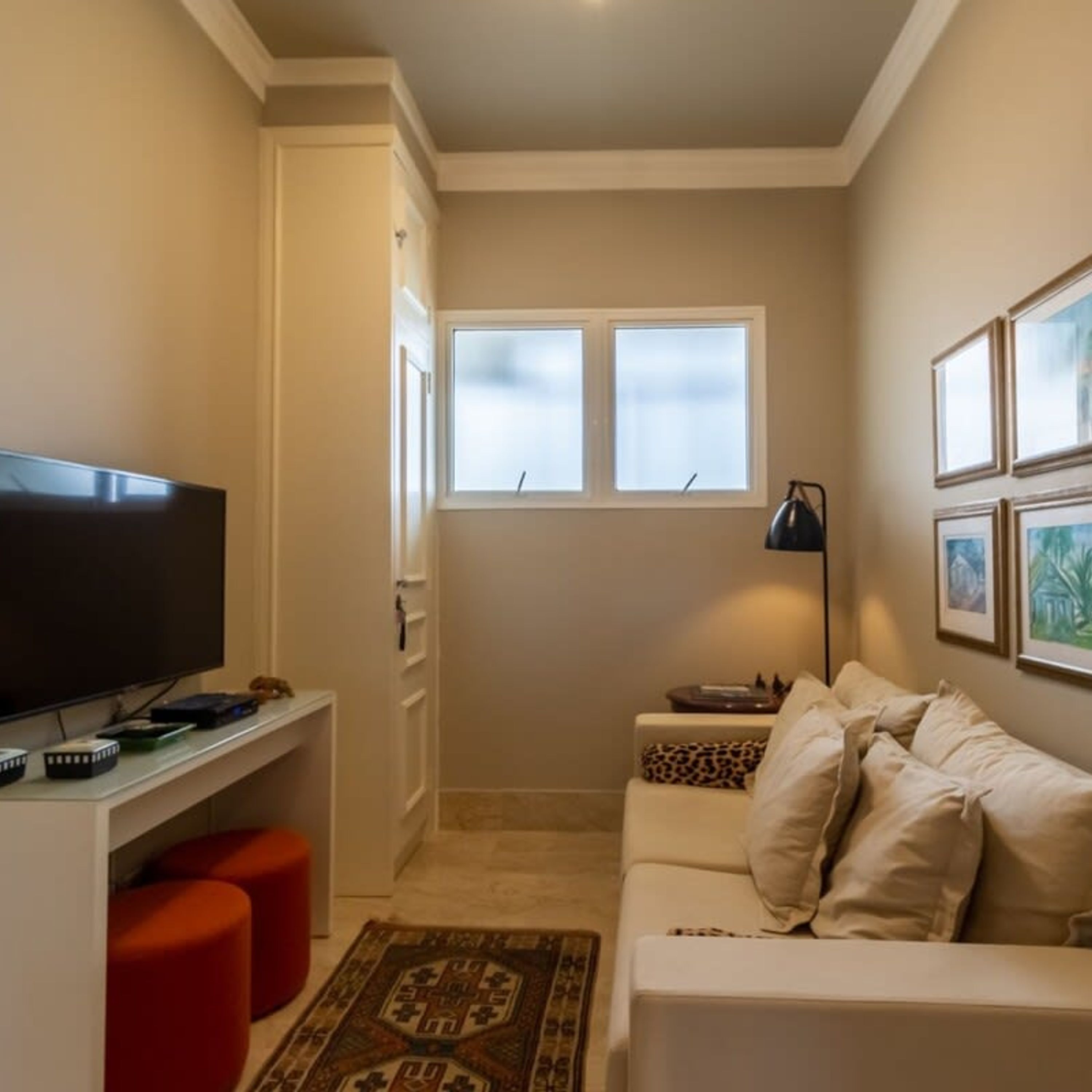 Un espace de vie compact comprenant un téléviseur à écran plat posé sur un meuble, un canapé blanc agrémenté de plusieurs coussins, un pouf rouge, un tapis à motifs et une fenêtre aux vitres dépolies.