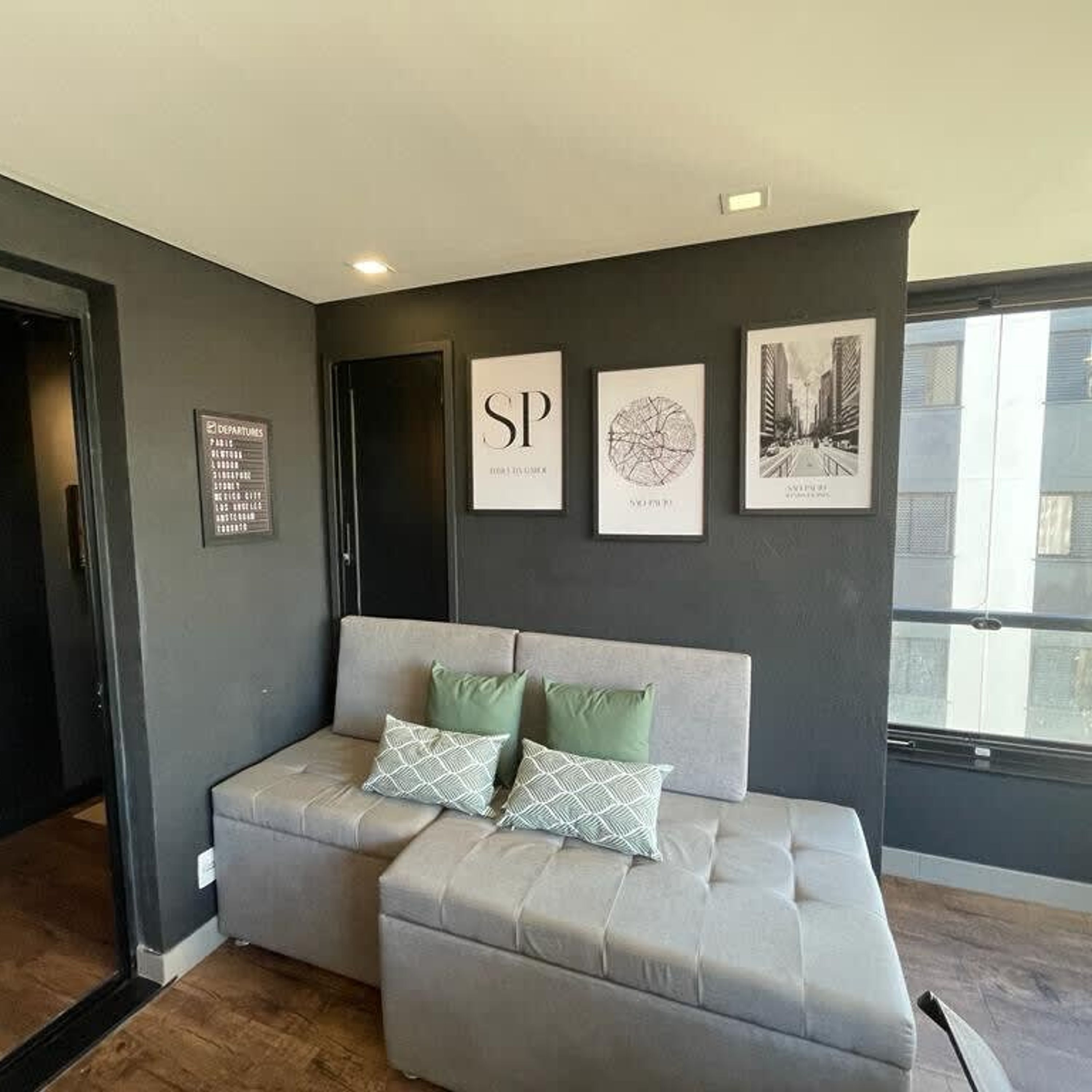 A modern living room with a grey sofa and ottoman, a wall-mounted sign listing departure cities, and three framed artworks above the sofa.