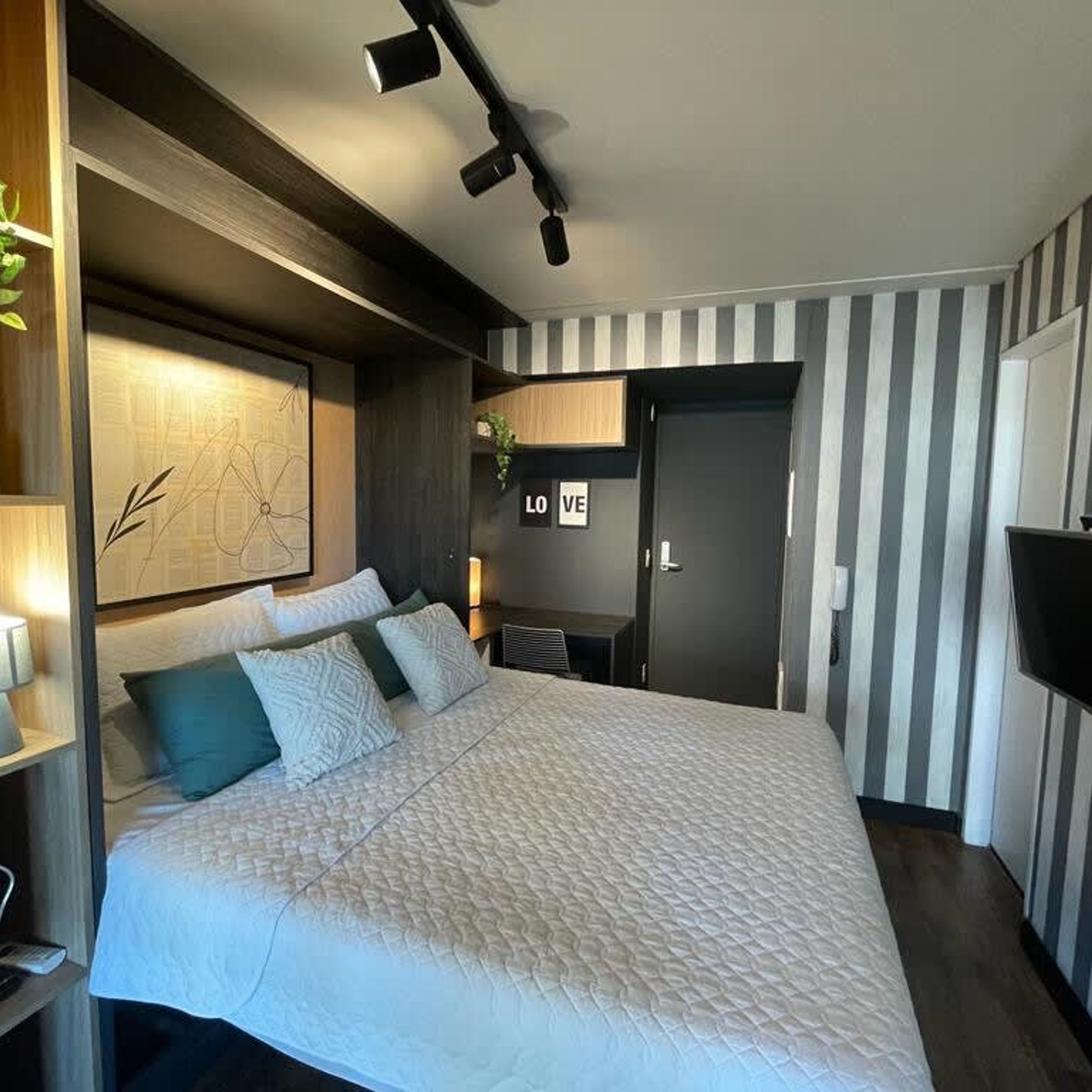 A modern hotel room with a large bed, a map artwork on the wall, a door with 'LOVE' sign, and a striped wall.