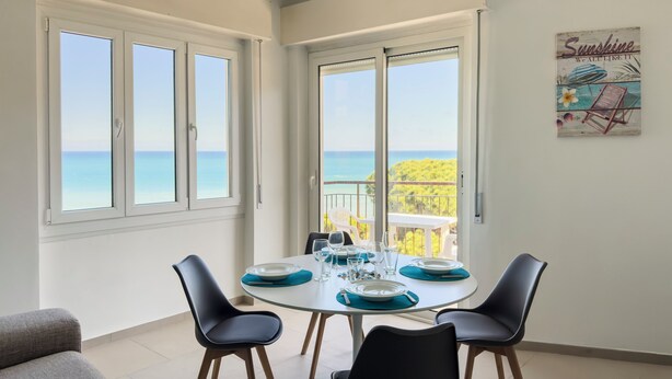 Un comedor con una mesa preparada para cuatro, ventanas amplias con vista al mar y un balcón con una silla.