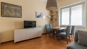 A living room with a TV on a white cabinet, a dining table with chairs, a bookshelf, and a large window.