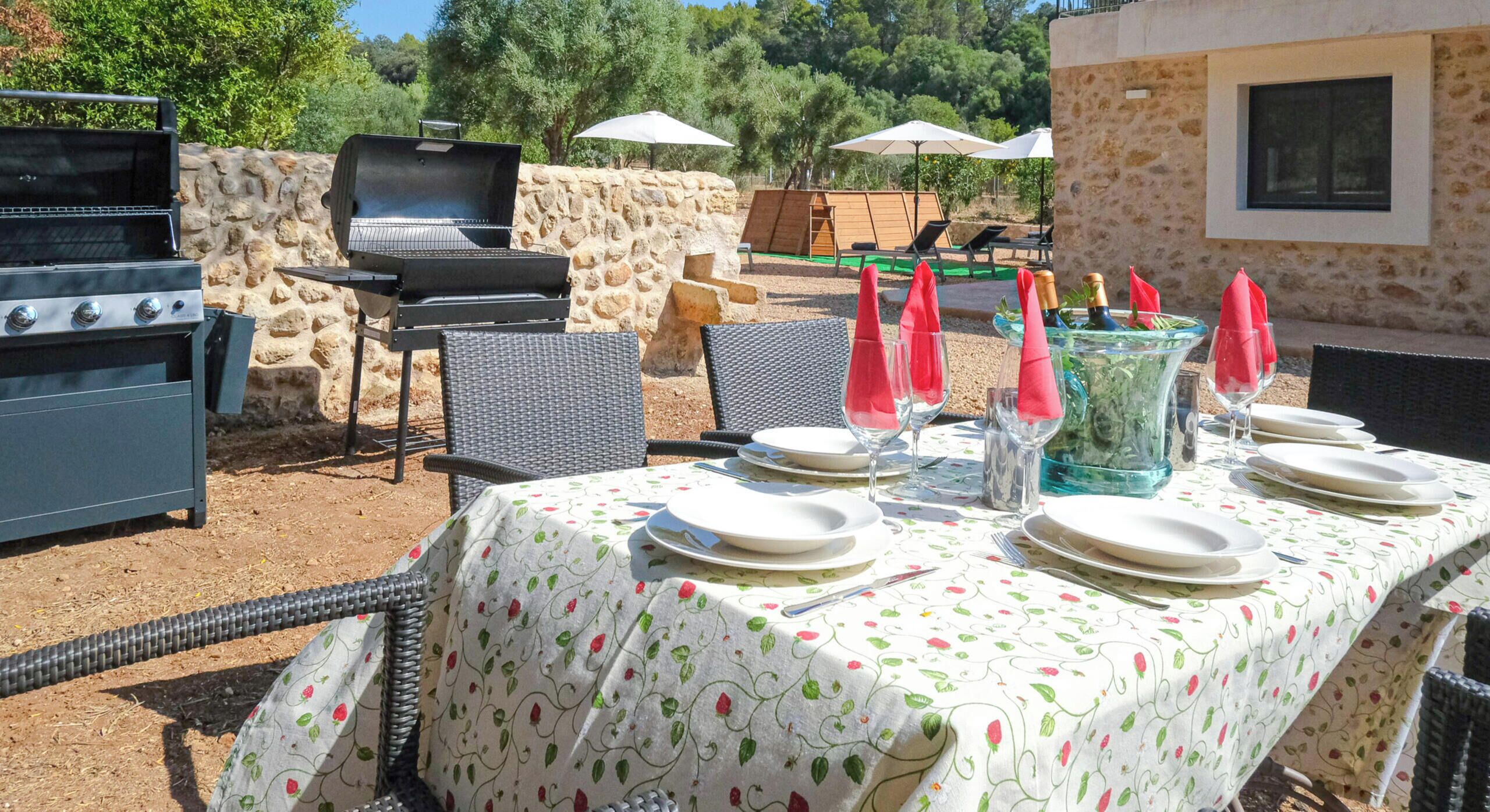 An outdoor dining area with a stone wall, a grill, and a table set for two with plates, cutlery, and red napkins.