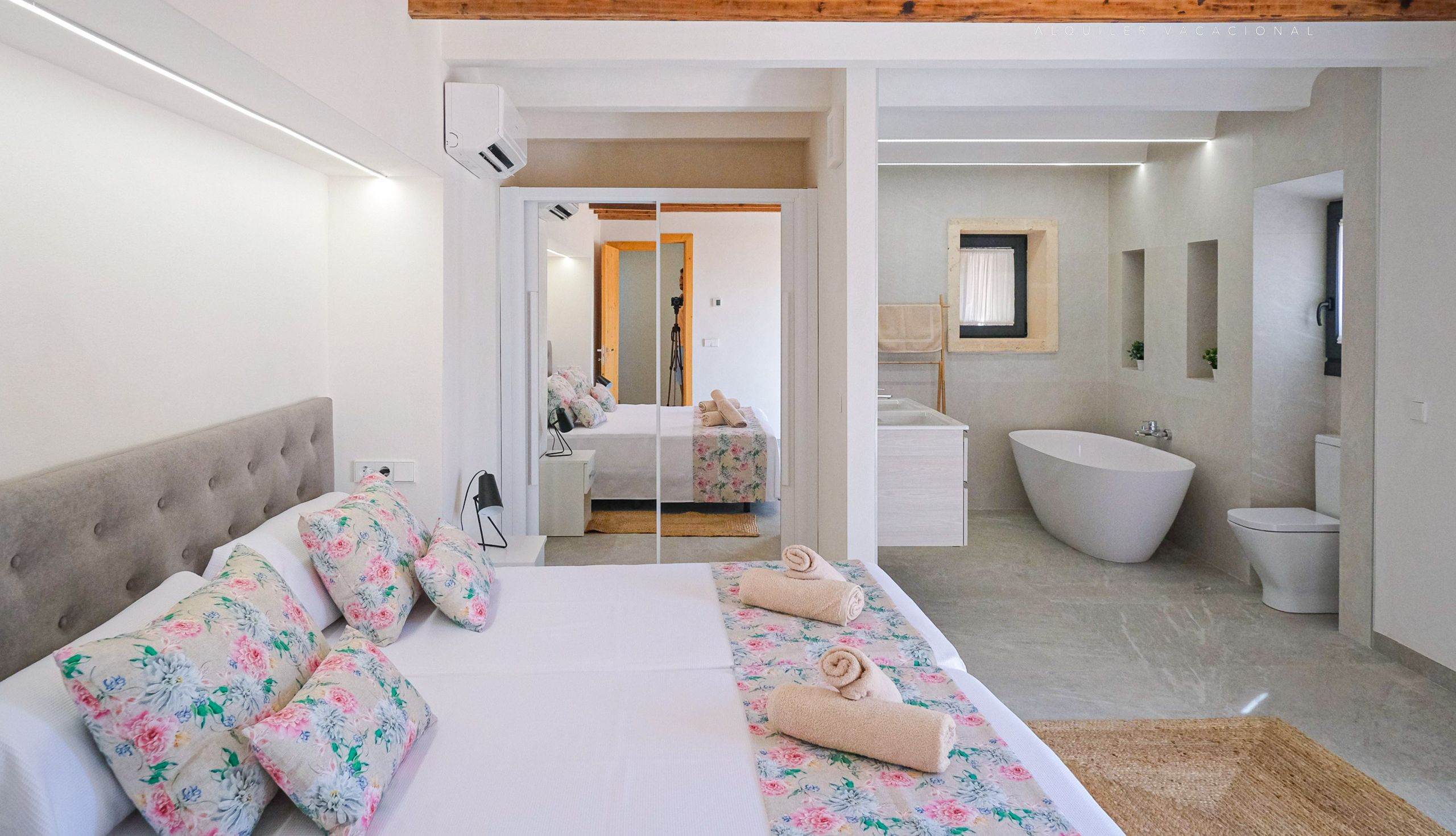 A modern bedroom with a large bed, floral pillows, a headboard, a bathroom with a bathtub, and a mirror.
