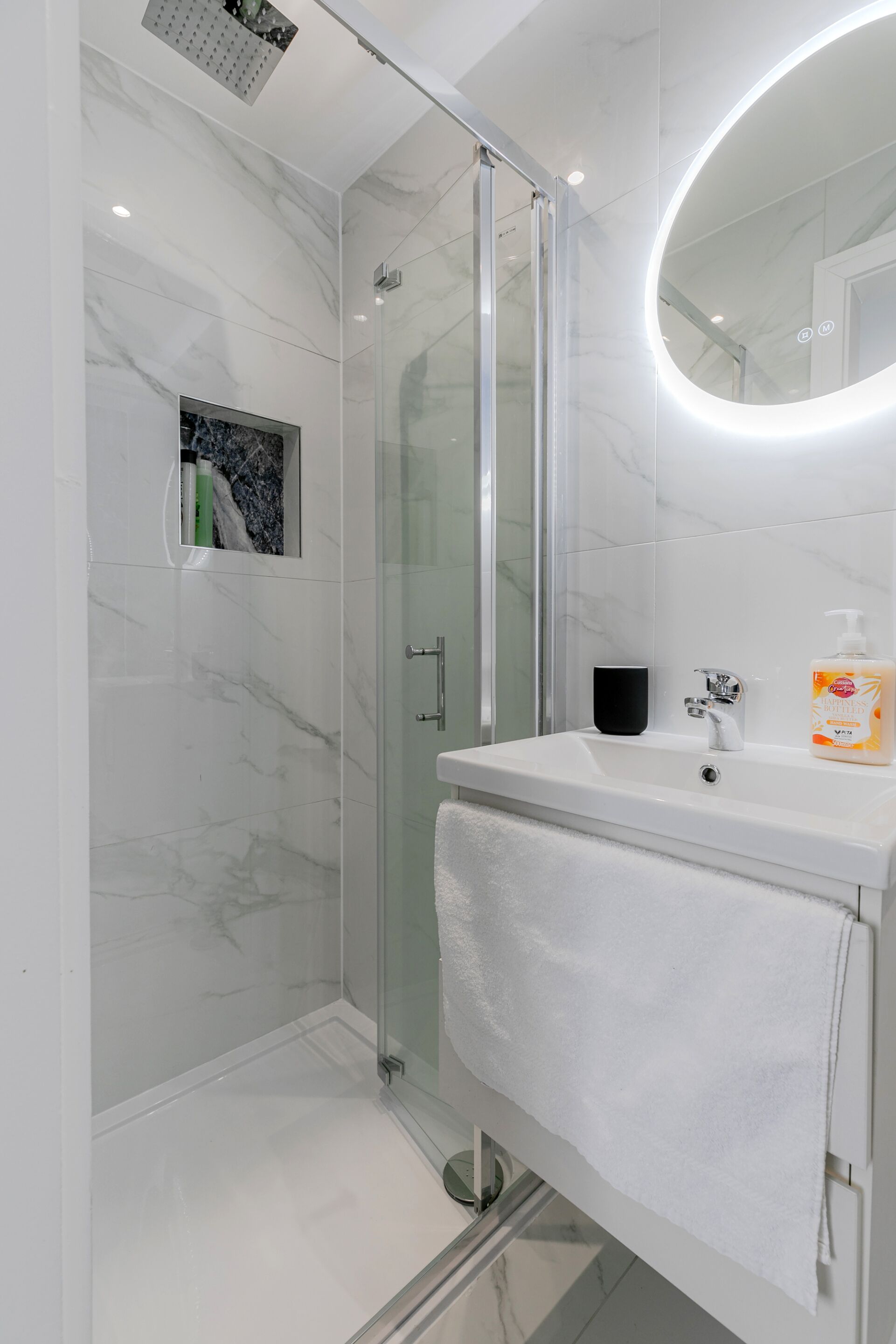 A modern bathroom with a glass shower enclosure, a white vanity with a round mirror, and a towel hanging on the wall.