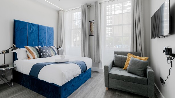 Una habitación de hotel moderna con una cama grande, un sofá gris, un televisor de pantalla plana y una ventana con cortinas.