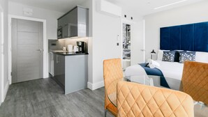Una habitación de hotel moderna con kitchenette, una mesa de comedor de cristal y una cama con cabecero azul.