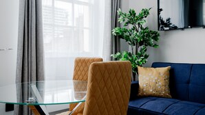 Una sala de estar moderna con una mesa de comedor de vidrio, dos sillas, un sofá azul, una planta y un televisor de pantalla plana.